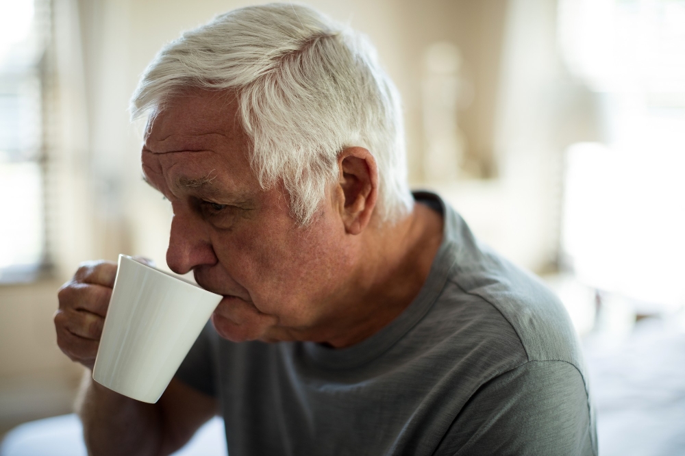Older man with white hair drinking his dysphagia diet drink from a white mug indoors